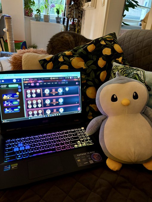 An MSI gaming laptop with an RGB keyboard displays what appears to be a Pokémon community page, sitting next to a plush penguin toy on a couch with lemon-patterned pillows, while a real cat sleeps in the background.