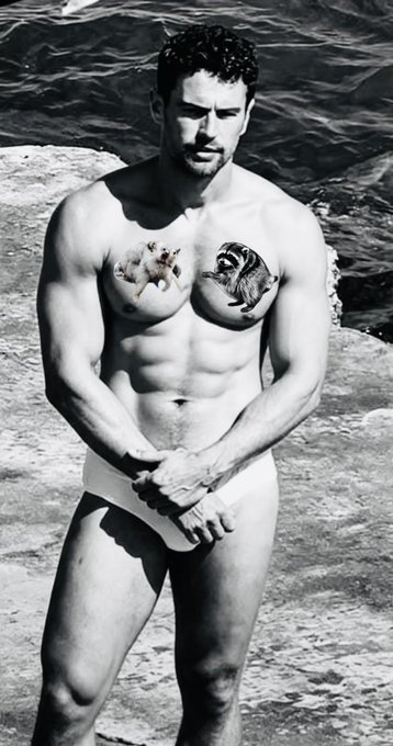 A black-and-white photo of a muscular shirtless man wearing white briefs on rocky terrain, with a cat tattooed on one nipple and what appears to be a ram skull tattoo on the other.