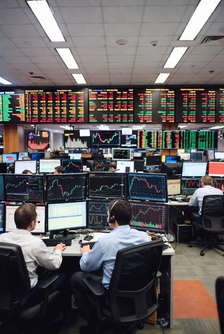 Two traders in dress shirts monitor multiple computer screens displaying financial charts while large digital boards above them show real-time stock market data with green and red figures.