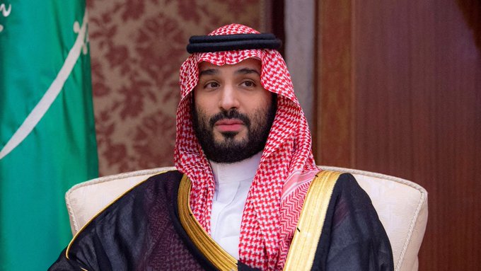 Saudi Crown Prince Mohammed bin Salman, shown seated in traditional Saudi attire beside the Saudi flag, as the kingdom faces a sharp drop in oil production to 6.2 million barrels per day due to the Iran war.
