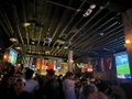 The image depicts a lively scene at a bar or pub where people are gathered around a TV screen, watching a football game. The room has a dark ceiling with exposed beams, giving it an industrial feel. There are numerous people present, some standing closer to the TV while others are scattered throughout the space. A few individuals can be seen holding drinks in their hands, enjoying the atmosphere and the company of others. The overall vibe suggests that this is a social event where friends have come together to watch sports or engage in conversation.
