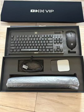 An 'OKX VIP' gift box set is opened to reveal a black keyboard, gaming mouse, cables, and a mousepad on a wooden floor.