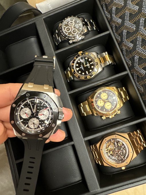 A hand holds a large black-and-silver chronograph watch over a display box filled with several gold and two-tone luxury-style watches. The post celebrates receiving the watches from TCG on Pumpfun and thanks @a1lon9.