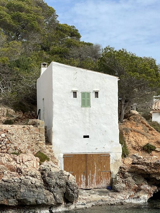A small, weathered whitewashed building with green shutters and a wooden garage door sits on rocky terrain beside the water, surrounded by lush green trees under a partly cloudy sky.