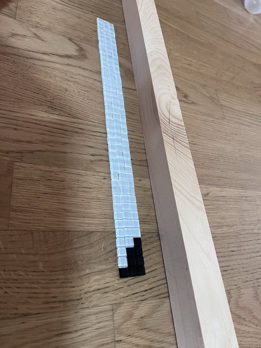 A long strip of white square tiles laid out on a wooden floor next to a long piece of light-colored wood, with a small section of black tiles at the end of the strip.