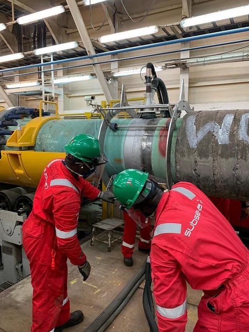 Three workers in red Subsea7 coveralls and green hard hats work on a large cylindrical industrial pipe inside a workshop.