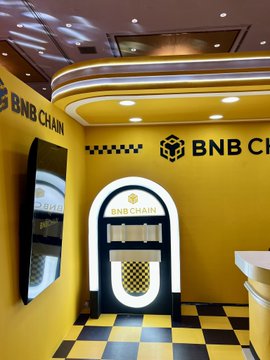 A brightly lit, yellow-walled room displays the 'BNB CHAIN' logo prominently above an arched jukebox-style kiosk and on the adjacent wall, complemented by a black and yellow checkered floor.