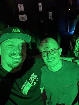 Two smiling men pose for a photo in a dark setting illuminated by green light, with small white papers stuck to the wall behind them.