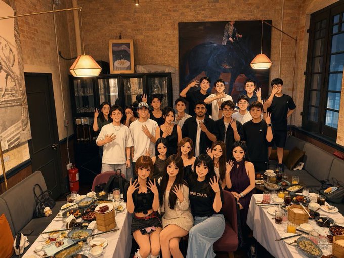 A large group of young people pose together at a dinner table, with some wearing shirts reading "BIN CHAN" and "BINANCIAN," celebrating their connection to the Binance community in a stylish, brick-walled restaurant with industrial lighting and a large elephant painting in the background.