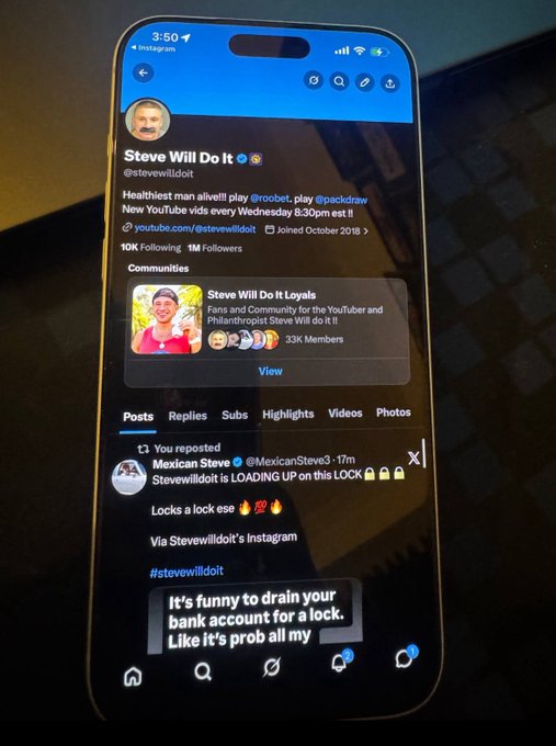 The image shows a cell phone with a Twitter app open, displaying a message from Steve Will It. The message reads "It's funny to drain your bank account for a lock. Like's all my prob." This message seems to be related to a recent incident where someone was locked out of their account and had to drain the account to get in.

The phone is placed on top of a table, with its screen facing upwards towards the viewer. The background appears to be a dark surface or a windowed area, providing contrast for the image.