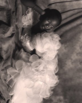 A black-and-white photograph of a person reclining in a flowing, ruffled gown with swirling translucent fabric around them.