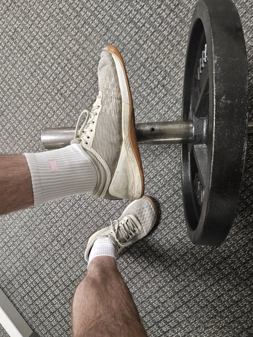 A first-person view looking down at a person's feet in white socks and worn white sneakers resting on a barbell with black weight plates on a rubber gym floor.