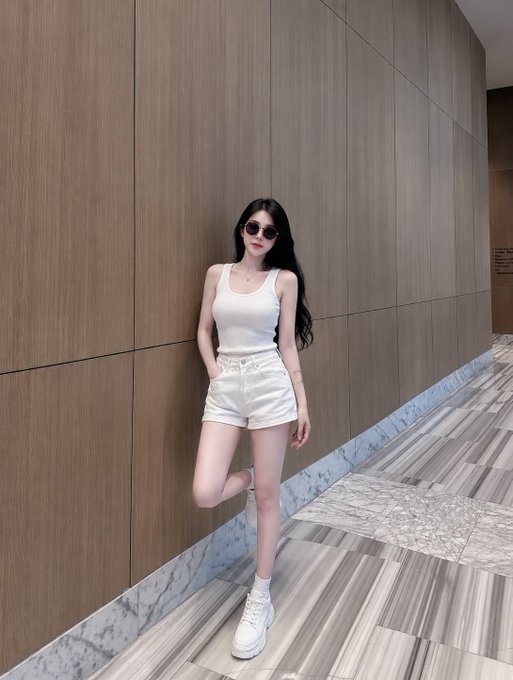 A woman wearing a white tank top, white denim shorts, white platform sneakers, and sunglasses, posing against a wood-paneled wall in a marble-floored hallway.