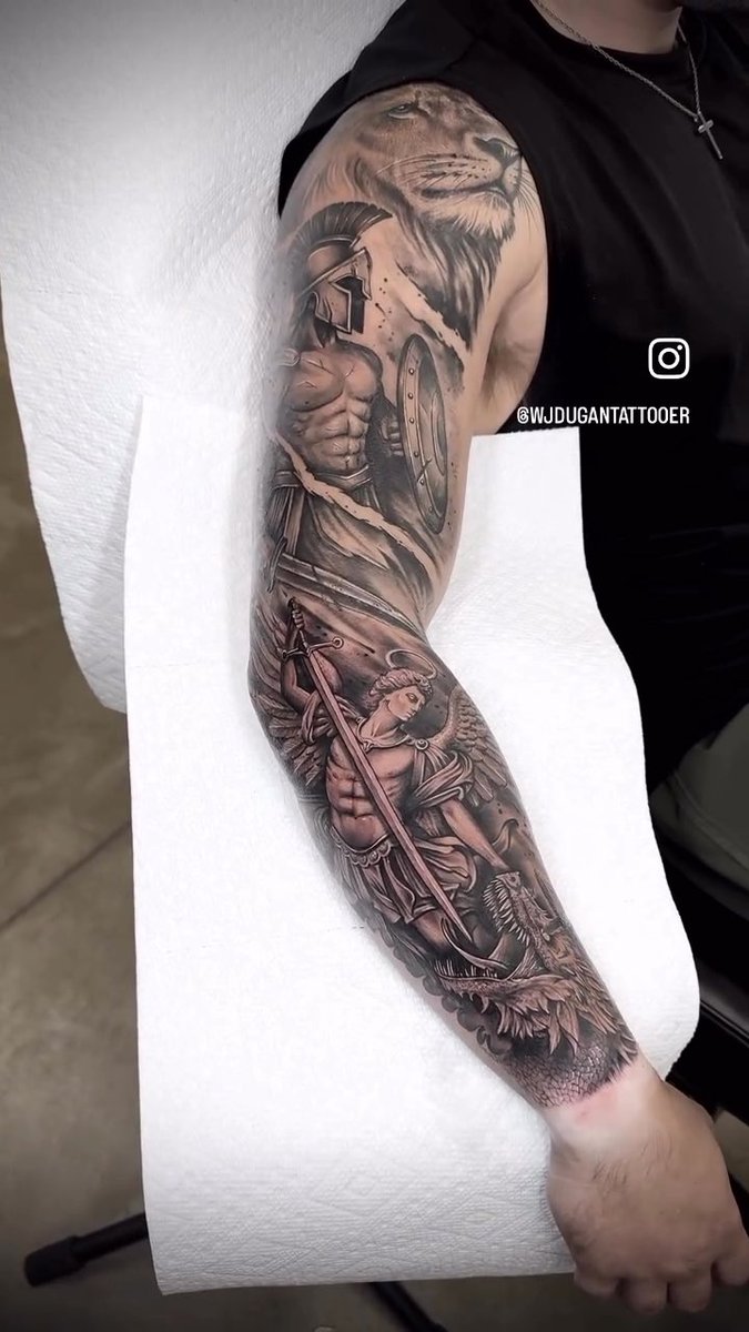 A detailed black-and-grey tattoo sleeve featuring a lion at the shoulder, a Spartan warrior with shield in the middle, and Archangel Michael with a sword and wings near the wrist, credited to @wjdugantattooer.