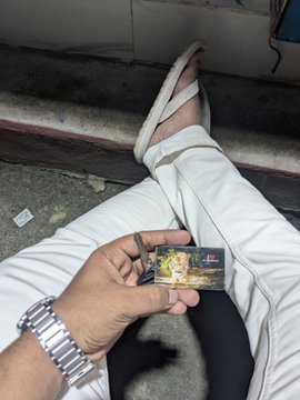A person holding a small Game Boy Advance SP cartridge outdoors, wearing white pants and a silver watch, with feet in sandals visible in the background.