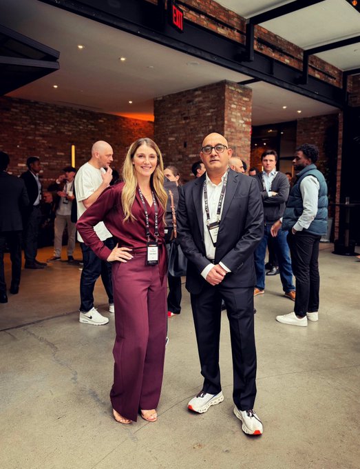 A woman in a burgundy jumpsuit and a man in a dark suit pose together in an industrial-style event space with exposed brick and other attendees in the background.