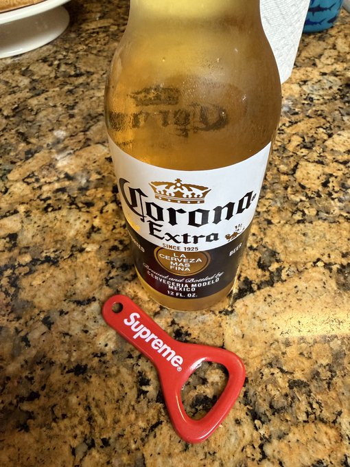 A Corona Extra beer bottle with a lime next to a red Supreme bottle opener on a granite countertop.
