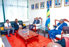 An indoor meeting in an official office with a Tanzanian flag in the background, where a group of men in suits are seated on sofas around a coffee table engaged in discussion.