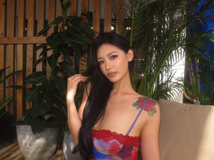 A young woman with long dark hair wearing a floral top poses in front of green plants, displaying a red rose tattoo on her left shoulder.