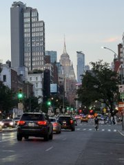 A street-level view looking down a city road toward the Empire State Building in the distance, featuring traffic with cars, traffic lights, and urban buildings on both sides.