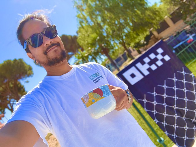 A man in sunglasses and a white "OFF•" t-shirt takes a selfie while holding a colorful cup, standing in front of an "OFF•" branded backdrop with checkered patterns. The "GM" post aligns with the sunny, cheerful vibe of his casual morning selfie in the Off-White branded setting.