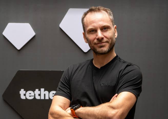 A smiling man with short brown hair stands with crossed arms in front of a dark wall decorated with white hexagonal panels and the "tether" logo, wearing a black T-shirt also branded with the Tether logo. The image visually reinforces the post by pairing Tether's branding with a confident, approachable spokesperson-style portrait, suggesting leadership behind the trend of crypto profits seeking opportunities elsewhere.