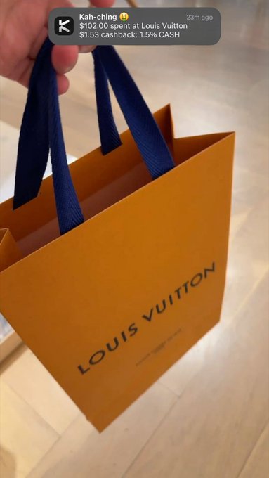The image shows a person holding an orange Louis Vuitton shopping bag over their shoulder. The bag has a blue ribbon handle and displays the brand name "Louis Vuitton" prominently. The person appears to be taking a photo of themselves with the bag, possibly sharing it on social media or sending it as a message.