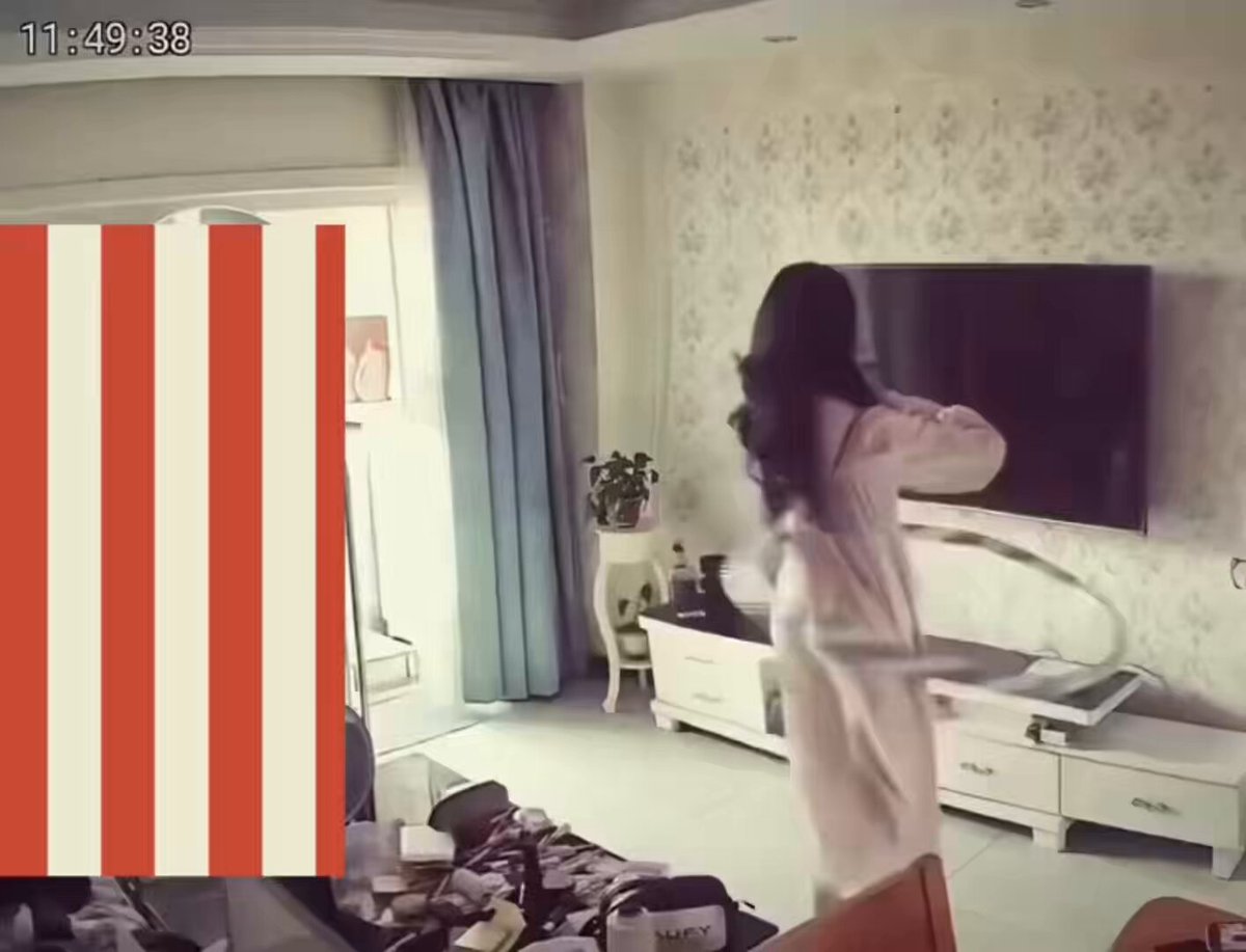 The image shows a woman standing in a living room, wearing a white dress with her hair styled in loose waves. She appears to be dancing or moving around the room while holding a plate of food. The room has a TV mounted on the wall above a couch and chairs arranged for seating. There is also a potted plant placed near one of the chairs.