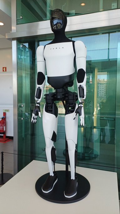 The image features a large, white and black robot standing upright on a pedestal or base. The robot appears to be made of metal and has a futuristic design with arms and legs that resemble human-like limbs. It's positioned near a glass window, which allows for an unobstructed view of the surroundings outside. In the background, there is another person who seems to be observing the robot closely.