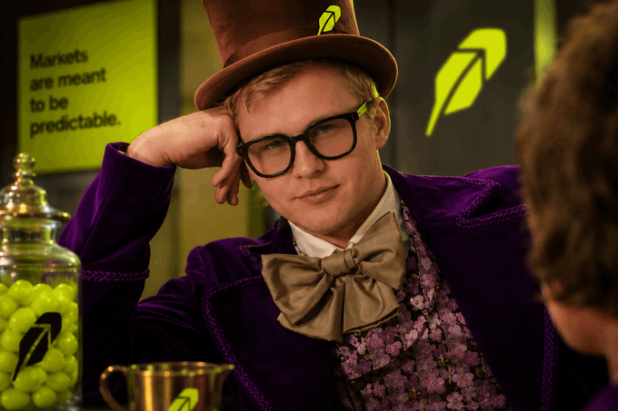 A bespectacled man in a purple velvet suit and brown top hat poses with a jar of green candies, alongside a sign reading "Markets are meant to be predictable," evoking a Willy Wonka-style lure to contrast with the post's warning that most new memecoins are rug pulls or failures, with only 1–2 breaking above a $1M market cap.