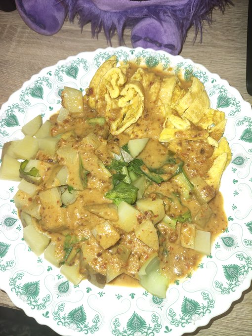 A plate of food on a decorative white and green patterned dish containing what appears to be a curry or stir-fry with egg, potatoes, cucumbers, and herbs, with purple fuzzy slippers visible in the background.