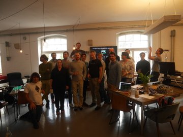 A group of around 15 casually dressed people pose together in a modern office space with large windows, desks, and computer equipment, with one person raising their arm in celebration. The scene connects to the caption by depicting the "smartest people" team behind the cryptocurrency and financial technology venture referenced in the post.