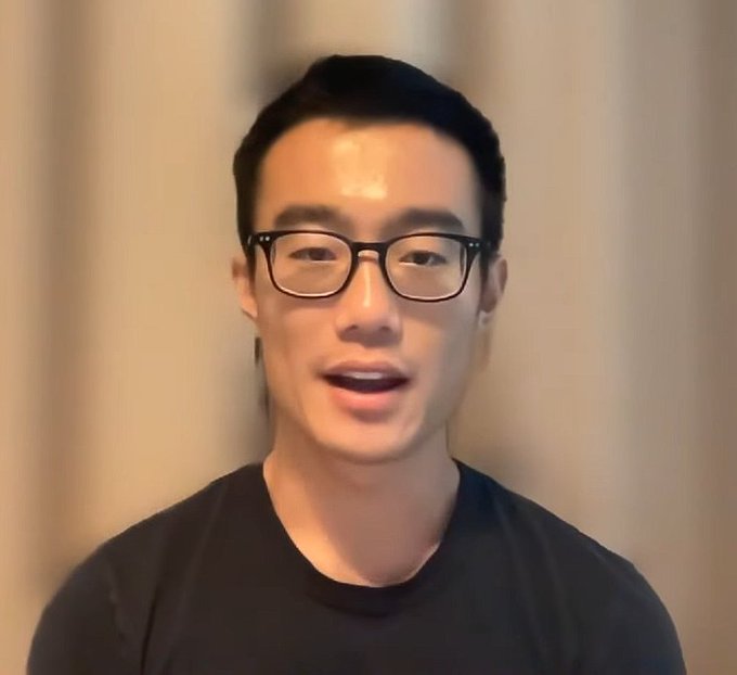 A close-up of an Asian man with short black hair and glasses wearing a black t-shirt, speaking against a blurred beige curtain background.