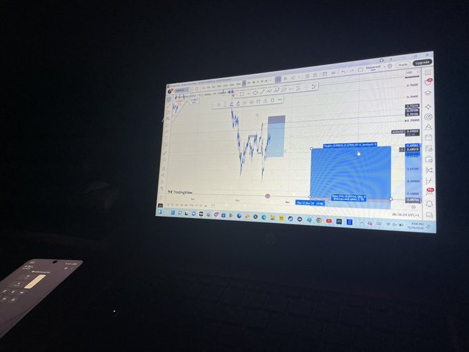 A laptop screen displaying a TradingView candlestick chart in a dark room, showing trading data with a phone resting beside it.