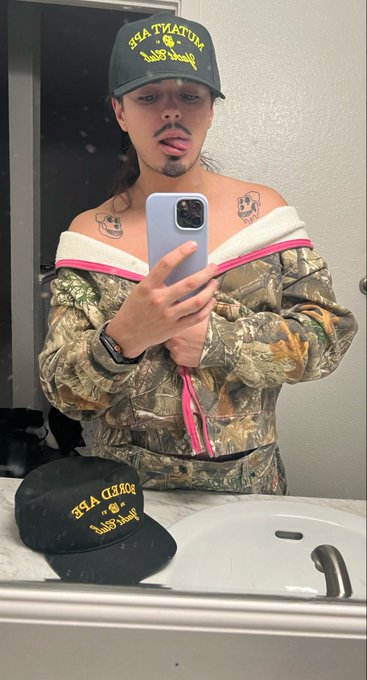 A selfie mirror photo of a person wearing a black 'SEA TIATUM' cap and an off-shoulder camo top with pink trim, with a matching cap sitting on the bathroom counter.