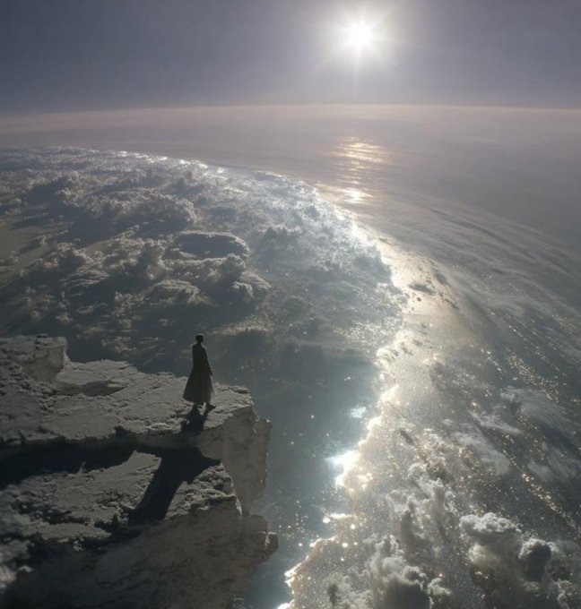 A lone figure in a long coat stands on a rocky cliff edge overlooking a vast, sun-reflecting expanse of water and clouds.