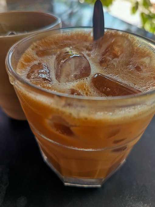 A close-up of an iced Thai tea or coffee in a clear glass filled with ice cubes and topped with foam, served next to a ceramic cup.