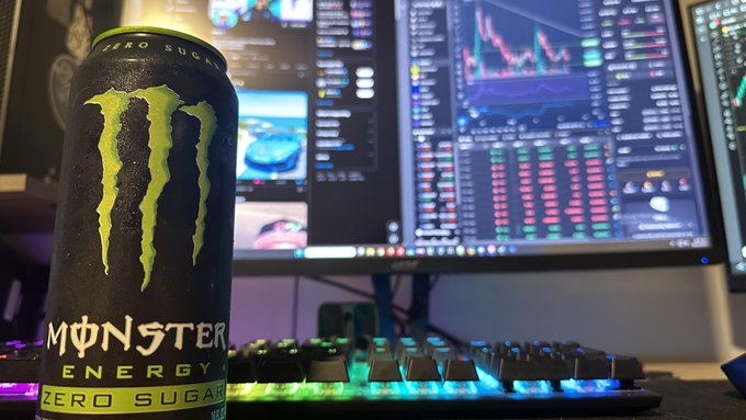 A can of 'MONSTER ENERGY ZERO SUGAR' sitting in front of a backlit RGB gaming keyboard and a monitor displaying crypto trading charts.