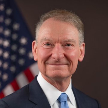 An official portrait of a man in a dark suit and light blue tie, smiling with an American flag visible in the background.