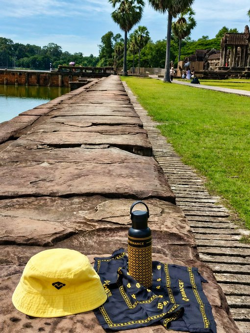 A yellow bucket hat and a black 'BINANCE' branded water bottle and towel are placed on a stone walkway next to a body of water with palm trees and a temple structure in the background.