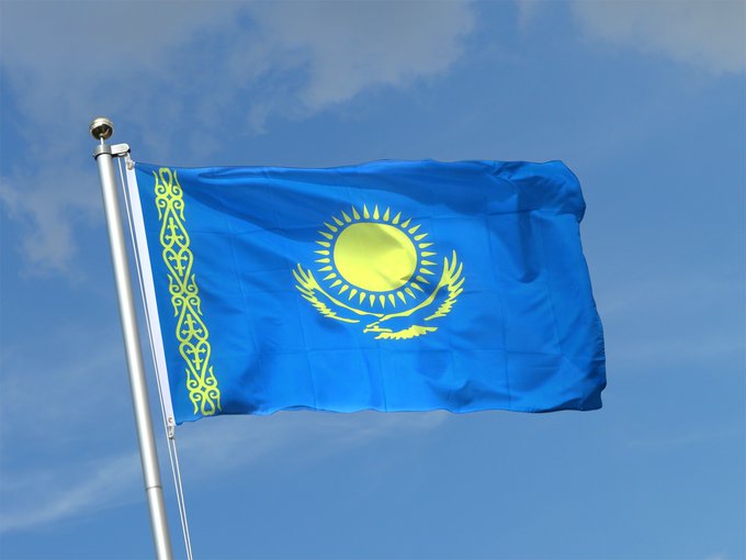 The national flag of Kazakhstan, featuring a sky-blue background with a golden sun and soaring eagle, alongside a vertical ornamental band on the hoist side, waving against a partly cloudy sky.