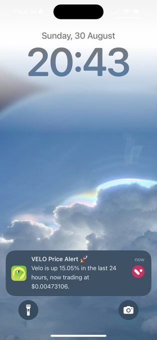An iPhone lock screen displays the time 20:43 on Sunday, 30 August against a stunning wallpaper of clouds with a rainbow halo. A "VELO Price Alert" notification appears, showing that Velo is up 15.05% in the last 24 hours, now trading at $0.00473106, perfectly aligning with the accompanying post about the $VELO price alert from @veloprotocol.