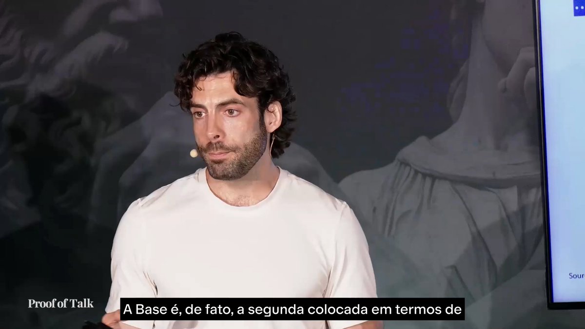 The image features a man with curly hair wearing a white shirt, standing in front of a large screen or television monitor that displays text related to cryptocurrency. The caption below the image reads "O que torna a tokenização tão importante é o que ela destrava: mercados de lending e borrowing em protocolos DeFi abertos." This suggests that the man is discussing the importance of cryptocurrency and its impact on the financial world, specifically focusing on the concept of DeFi.