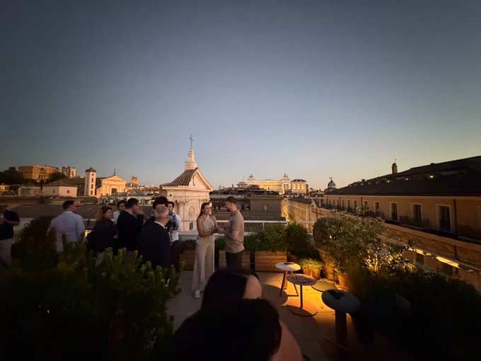 Guests mingle at a rooftop event in Rome during golden hour, with the city's historic skyline—including a cross-topped cupola and domed landmarks—stretching into the distance. The caption "YESTERDAY'S @cryptocom EVENT IN ROME!" reflects the elegant gathering shown, with attendees chatting near potted plants and bistro tables against a stunning panoramic view.