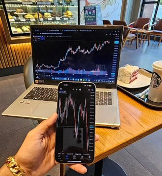 A hand holds a smartphone displaying a stock chart in front of a laptop showing a similar trading chart, with a slice of cake and a Starbucks coffee on the table in a café setting.