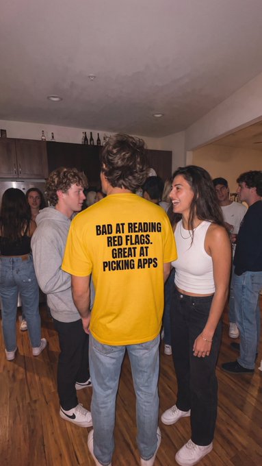 A young man in a yellow t-shirt reading "BAD AT READING RED FLAGS. GREAT AT PICKING APPS" chats with a smiling woman at a house party, cleverly promoting the Binance crypto app through the shirt's self-deprecating joke about spotting good "apps" (both dating and digital).