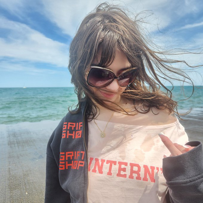 A young woman with windswept hair and cat-eye sunglasses poses at the beach, wearing a layered outfit featuring a jacket and shirt branded with "GRIFTSHOP" and "INTERN" text—showcasing the new Griftshop intern program swag mentioned in the post.