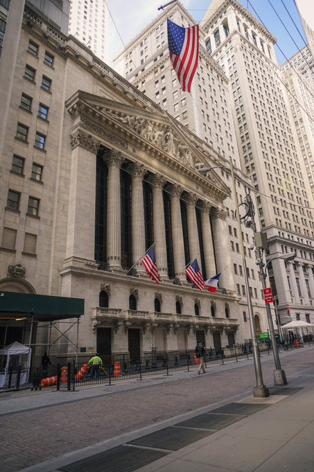 A street-level view of the New York Stock Exchange building, featuring its iconic columns and American flags, with pedestrians and workers visible on the sidewalk.