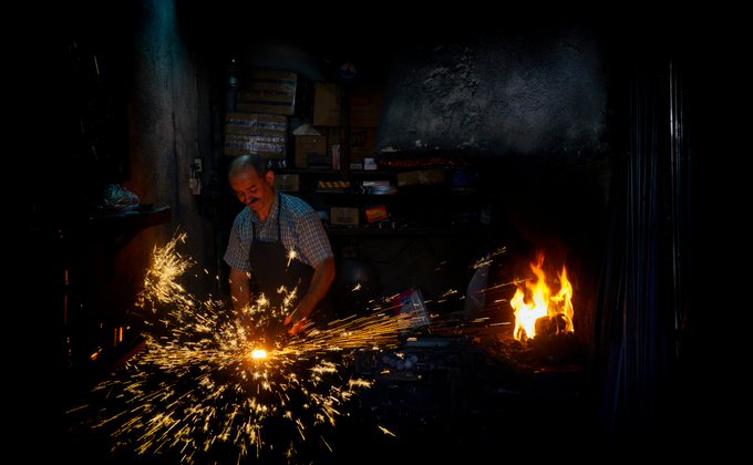 A blacksmith works at a forge in a dark workshop, with bright orange sparks flying as he shapes hot metal, illuminated by the glow of a fire nearby.