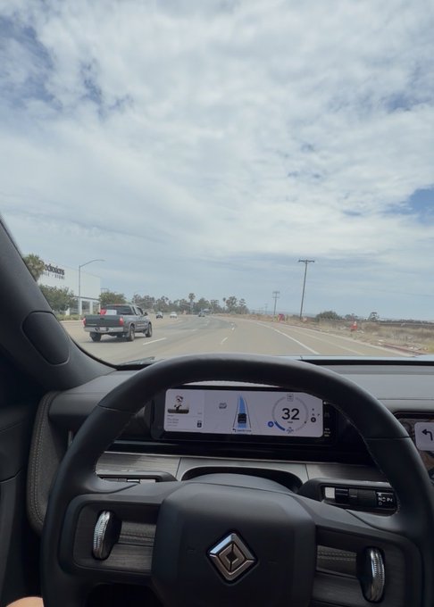 A view from the driver's seat of a vehicle showing the dashboard, steering wheel with a logo, and a digital speedometer reading 32 mph on a multi-lane highway.